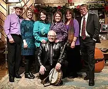 Seven white adults in semi-formal clothing are standing on a stage in front of a wooden sign that reads "Carter Fold".