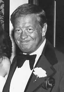 Black and white image of a man in a suit and bowtie, with a white flower pinned on his lapel