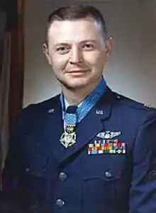 Head and shoulders of a man with short hair and a round face in a dark blue military jacket. Rows of ribbon bars and a winged pin are on his left breast and a medal hangs from a light blue ribbon around his neck.