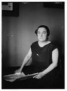 A white woman with dark hair, wearing a dark dress, seated indoors