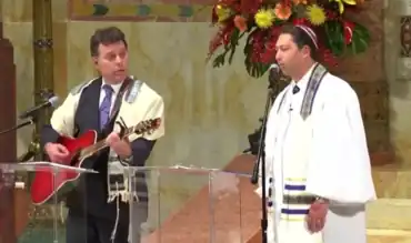 On the right, a cantor sings into a microphone. Standing to his left on the bimah (stage), another man sings along and strums a guitar. Both are wearing tallitot (prayer shawls) atop suits, and the cantor is visibly wearing a kippah (skullcap); the guitarist is positioned such that his kippah is not visible to the camera.