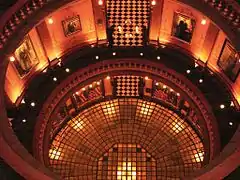 A cylindrical room with a glass floor surrounded by two higher stories of circular balconies with ornate bulbous balusters. The room is lit by incandescent lights.