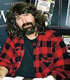 A male with medium-length hair and a goatee, wearing a red-and-black plaid buttoned shirt on top of a black T-shirt poses at an autograph signing.