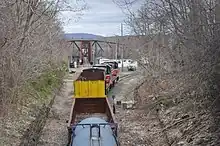 A P&W train is seen from a bridge which crosses over the track it is on. It is moving away from the viewer, and lead by two diesel locomotives, followed by several assorted freight cars. A railroad swing bridge can be seen in the distance.