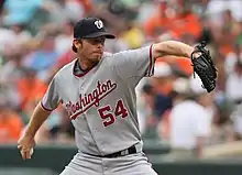 A man in a gray baseball uniform with "WASHINGTON 54" on the chest, a dark baseball cap, and a baseball glove on his left hand pitches a baseball with his right hand.