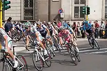 A large group of cyclists makes a right turn around a street corner in a major city. Prominent among them is a cyclist wearing an all-pink jersey, which represents being the overall leader in the race. A crowd of spectators watches the group of cyclists from the roadside.