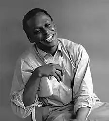 Miles Davis is seated and smiles while looking at the floor.