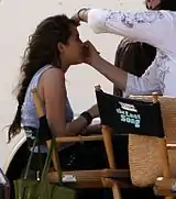 A side-view shot of a female teenager with long brown hair who smiles as a woman with dark brown hair, glasses, and a white shirt bends to brush the teen's face with make-up. The teen sits in a chair. Beside her is an identical chair with its back facing the camera; the chair's back has the words "The Last Song" written across it.