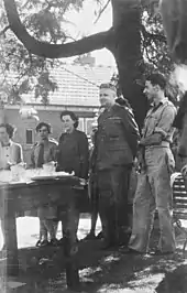 Black and white photograph of a group of three women and two men wearing military uniform. All members of the group are standing under a tree and are looking at a table.