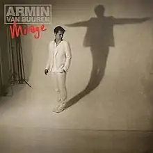 In a photo studio, Armin Van Buuren (in white clothing seen looking left) standing on a white background with a shadow of him seen with his arms extending outward.