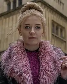 Woman with blonde hair in a bun looking at the camera, wearing a pink fluffy coat and a pink fluffy jumper