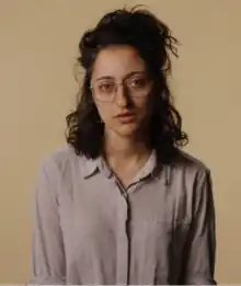 An image of Mitra Jouhari, who has curly brown hair that is half up and half down. She is wearing large square eyeglasses and looking directly at the camera. She has olive skin and light green eyes.