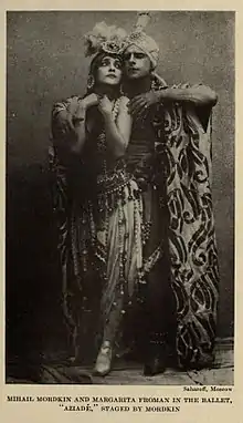 Two dancers; the male dancer behind the female dancer, with his one arm around her; they are wearing elaborately embellished costumes, and she has a large feather plume on her headdress.)