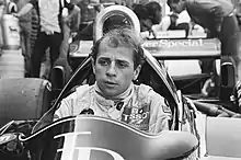 A black and white photograph of a man sitting inside a stationary racing car without a racing helmet but in racing overalls