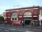 A red-bricked building with a rectangular, white sign reading "MORNINGTON CRESCENT STATION" in black letters all under a light blue sky