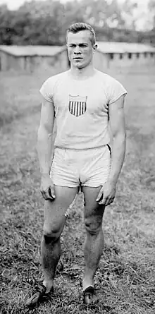 A young man wearing an all-white singlet and shorts stands in a grassy field.