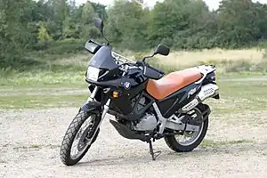 A black BMW F650 fitted with an orange seat, parked on an area of open ground