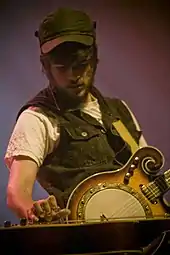 A man wearing a cap, denim jacket, and old guitar.