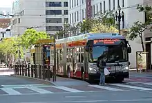 A silver bus at a boarding island on a wide street