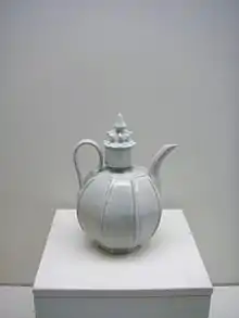 A white teapot with an almost perfectly spherical body and a large, cylindrical cap in the center which is topped with a small crown shaped embellishment. Several vertical lines are glazed into the body of the pot.