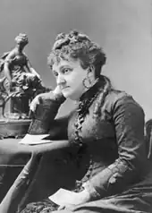 Black and white photograph of a middle age white woman sitting at a table with her left arm on her lap and her right arm on the table. She wears a dark dress and has long dark hair tied up on the top of her head. She has a faraway look on her face.