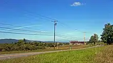 A two-lane highway traverses open fields in a rural area. In the background is a large ridge; a second ridge is barely visible in the distance.