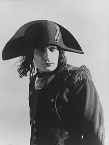 A monochrome photographic portrait of a handsome man in his late 20s wearing a French general's uniform from the 1790s and a cocked hat over stringy dark hair that reaches his shoulders