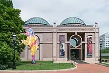 A one-story symmetrical granite building with two green domed roofs either side of center. The entrance door is on the right and a similar design is on the left made out of a slightly darker granite.