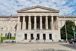 A 3-story building. The first story is granite, while the second two are sandstone. There is a pedimented front with columns spanning the second and third floors and the entrance on the first.