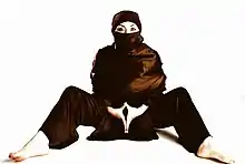 A woman wearing a hijab with black shirt and pants sits on the ground, with her hands pressed together