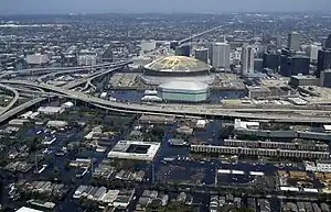 Aerial view of a downtown area of a city in the background and suburbs in the foreground inundated by floodwater. Except for the highways, nearly every street is completely covered in water. Various buildings, particularly the dome shaped building in the center, show damage.