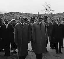 Two men standing side by side in the forefront, wearing overcoats. Behind them are several men in military uniform or suits and ties standing and saluting or making no gestures.