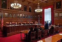 A room with ornate brown wooden paneling and oil portraits on the walls. At the left seven people wearing black robes sit behind a similarly decorated wooden bench, elevated slightly from the red-carpeted floor. On the right are several people in suits sitting at chairs behind tables. In the rear is a large window with red drapes.