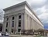 A long light-colored stone building seen from across the street. It has round fluted columns with ornate capitals running across its front, from one side of the image to the other.