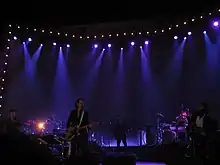 A rock band performing live on-stage. To the left, one man sings into a microphone and another man plays drums. In the centre, one man plays guitar and another man plays bass. To the right, one man plays a violin and another man plays drums.