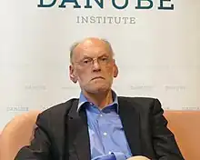 Nigel Biggar, wearing dark blazer and light blue shirt, sitting on a salmon-coloured sofa, looking directly at camera with serious gaze