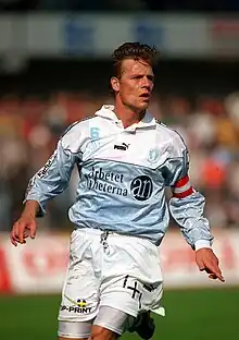 A dark haired man in light blue and white football attire looks to his left amidst a match. He wears a red and white striped armband on his left arm.