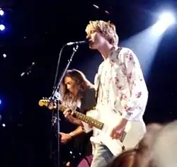 Kurt Cobain, in blonde hair, singing into a microphone onstage while Krist Novoselic plays bass in the background