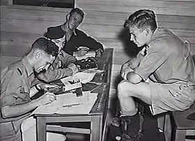 Three men writing at desks, while another man supervises