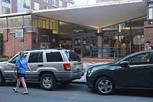 The outside of the one-story North End Branch of the Boston Public Library system. There are two cars parked in front, and a person in a blue t-shire and black shorts walking in the street. The building is mostly brick, but there is a section of doors and windows for the entrance.