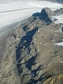 An aerial view of a sparsely snow-covered mountain slope with a glacier in the background.