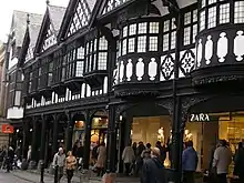 Part of a range of gabled shops seen from ground level. The lower storey contains modern shop fronts behind an arcade, The upper storey is timber-framed and contains bow windows in the foreground and oriel windows beyond.