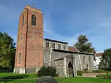 A church seen from the southwest with a large brick tower on the left and the flint body of the church on the right, showing the clerestory, south aisle and porch