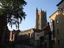 Protruding above the houses in a street is a battlemented tower with a corner turret, and the body of the church beyond it