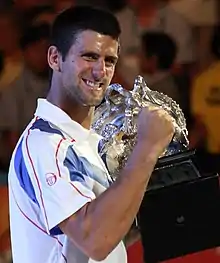 A brown-haired man in a white tennis shirt with light blue sections and red stripes with the trophy