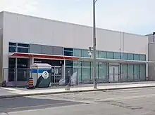 Photo of a light rail station entrance under construction, close to fully completed. It is a low slung building in white with glass panelling and an orange accent.