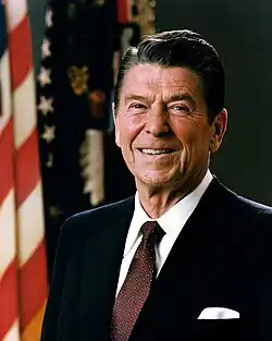 A photograph of a dark-haired, old man, wearing a suit and smiling in front of an American flag