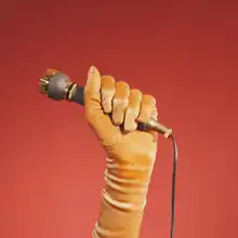 A yellow gloved hand holds a brass microphone amongst a red gradient background.