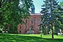 A two-story brown stone building with a green cupola and a statue in front, within a park