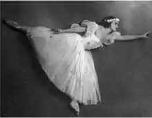 Photograph of a ballerina standing en pointe on her left foot with her right leg and torso extended perpendicular to the left leg. She is wearing a long tutu adorned with raised roses, which has a tight bodice with puffed sleeves, and a crown of flowers atop her head.